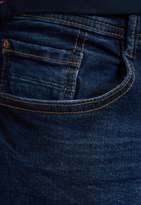 Jeans de mezclilla azul oscuro con costuras amarillas, que cuentan con un bolsillo frontal y una tela texturizada. La vista en primer plano enfatiza el material y los detalles de las costuras.