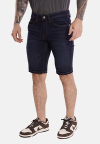 Shorts in denim blu scuro con una vestibilità slim, orlo arrotolato e cuciture visibili. Abbinati a sneakers marroni e bianche.