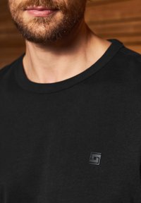 Czarna bawełniana koszulka z okrągłym dekoltem i małym, wytłoczonym logo na klatce piersiowej. Teksturowana tkanina o gładkim wykończeniu.