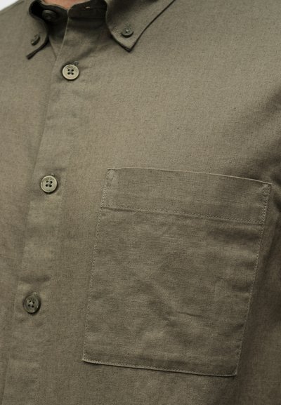 Gros plan d'une chemise kaki à boutons montrant la poche poitrine, trois boutons et une partie du col.