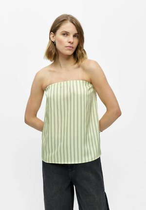Jeune femme aux cheveux mi-longs portant un top jaune pâle sans bretelles avec des rayures noires verticales et un pantalon ample foncé sur fond blanc.