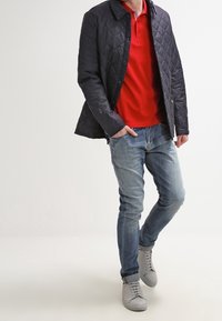 Mann in einer marineblauen Steppjacke über einem roten Poloshirt, mit ausgewaschenen blauen Jeans und hellgrauen Turnschuhen, der mit einer Hand in der Tasche steht.