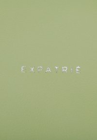 Green textured leather with embossed silver lettering reading "EXPATRIÉ". The surface is smooth with a slight sheen. No additional details visible.