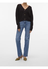 Musta neulottu lyhyt neuletakki V-aukkolla ja kolmella isolla napilla, yhdistetty suoralahkeisiin sinisiin farkkuihin ja oliivinvihreisiin kärjestä teräviin korkokenkiin.