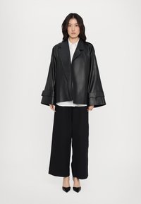 Jachetă oversized din piele neagră, cu mâneci largi, asortată cu o cămașă albă și pantaloni largi negri. Dispune de un design curat, minimalist.