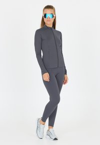 Grå atletisk jakke med høj krave og lynlås, parret med matchende leggings. Reflekterende blå solbriller og lette løbesko.