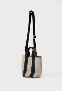 Μικρή μπεζ τσάντα από ύφασμα με μαύρους ραμμένους χειρολαβές και ρυθμιζόμενο ιμάντα ώμου σε απλό ανοιχτό φόντο.