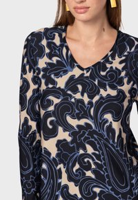 Top a maniche lunghe con scollatura a V in tessuto beige con un grande motivo floreale blu scuro, caratterizzato da dettagli blu e una texture liscia.