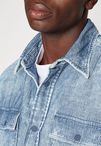 Gros plan d'une personne portant une veste en jean rayée bleue et blanche sur une chemise blanche, mettant l'accent sur le col et la poitrine.