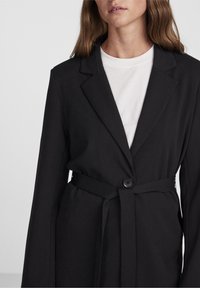 Černý blazer s upraveným designem, který má zapínání na jeden knoflík a odpovídající pásek v pase. Hladká tkanina a klopové límce.