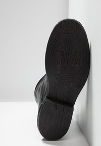Suela de bota de cuero negro con patrón texturizado, que presenta detalles de marca y modelo en relieve, en un ángulo sobre un fondo gris claro.