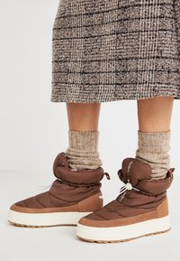 Botas tobilleras hinchadas de color marrón con acabado texturizado, suela de goma blanca y parte superior ajustable; combinadas con calcetines beige acanalados.