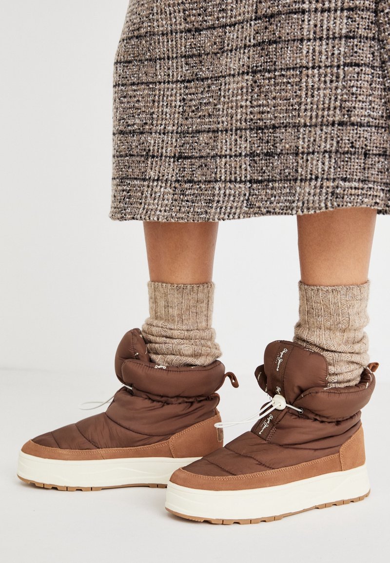 Botas tobilleras hinchadas de color marrón con acabado texturizado, suela de goma blanca y parte superior ajustable; combinadas con calcetines beige acanalados.