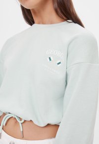 Světle zelená mikina s dlouhými rukávy, se zvýrazněným logem zkřížených vlajek na přední straně. Šňůrka na spodním lemu pro nastavitelnou velikost. Měkká textura.