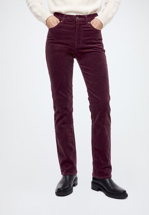 Le modèle porte un jean bordeaux à jambes droites, un pull blanc et des bottines noires, les mains dans les poches avant, sur un fond de studio uni.