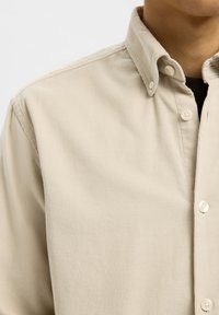 Beige ribfluwelen overhemd met knopen, gedragen over een zwart ondershirt, met zicht op de kraag en het bovenste deel van de borst.