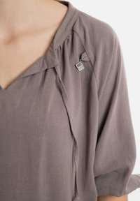 Lichtgrijze blouse met een V-hals en losse mouwen. Heeft een klein zilveren label en zachte, gestructureerde stof met subtiele plooien bij de halslijn.