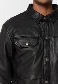 Giacca in pelle nera con collo, bottoni a scatto e due tasche sul petto. Texture liscia con un design aderente.