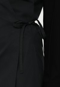 Čierny odev s viazaným pásom, ktorý ukazuje hladkú textúru a elegantnú siluetu. Bočný rozparok zdôrazňuje dizajn.