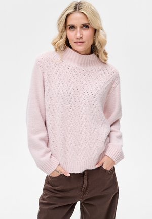 Różowy sweter dziergany na drutach z wysokim kołnierzem i wzorem w jodełkę. Posiada luźne rękawy i ściągacze przy mankietach. Miękka tekstura. Modelka nosi brązowe spodnie.