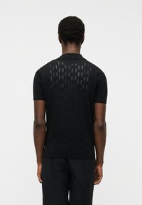 Άνδρας που φοράει μαύρο κοντομάνικο πόλο μπλουζάκι με διακριτικό διάτρητο μοτίβο στην πλάτη, συνδυασμένο με μαύρο σορτς, στέκεται μπροστά σε απλό λευκό φόντο.