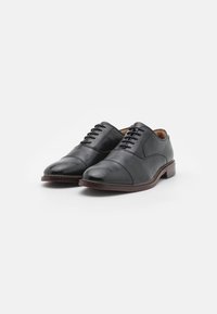 Chaussures habillées en cuir noir avec une finition lisse, bout rond, six paires d'œillets et semelle brun contrastante. Design classique adapté à une tenue formelle.