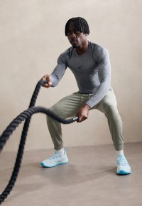 Athlete using a black battle rope in a workout, wearing a grey long-sleeve Nike shirt, light green sweatpants, and blue shoes.