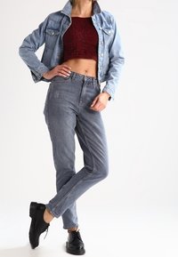 Žena pózuje v krátkém bordó krajkovém topu, světle modré džínové bundě, šedých kalhotách s vysokým pasem a černých šněrovacích botách.