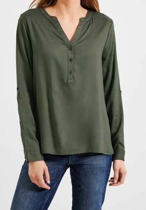 Femme portant un chemisier à manches longues vert olive avec un col en V et trois boutons, associé à un jean en denim bleu, sur fond blanc.