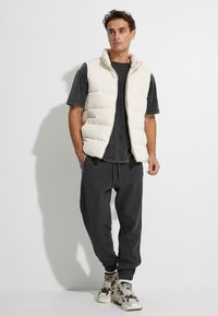 Helle, gepolsterte Weste, graues T-Shirt, dunkelgraue Jogginghosen und gemusterte Sneakers. Ärmelloses Design mit hohem Kragen und Seitentaschen.