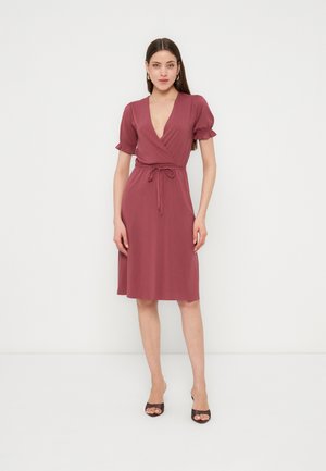 Kvinne iført knelang støvet rosa omslagskjole med puffede korte ermer, knyttet midje og svarte åpen tå-hæler, stående foran en ensfarget hvit bakgrunn.