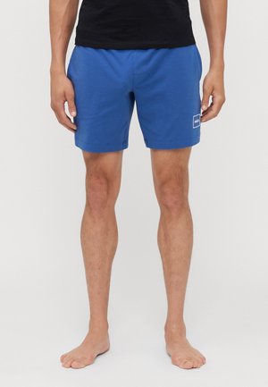 Shorts en coton bleu avec une taille élastique, coupe standard et un petit patch logo blanc sur la hanche gauche. Texture lisse.