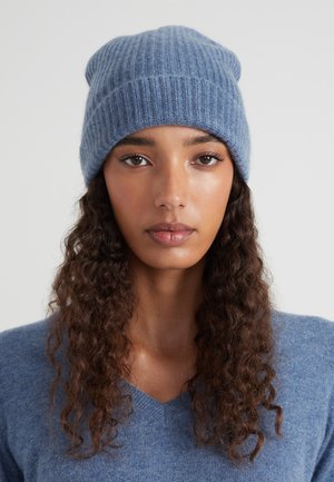 Junge Frau mit langen, lockigen Haaren, die eine blaue Strickmütze und einen passenden blauen V-Ausschnitt-Pullover trägt, neutraler Hintergrund.