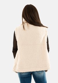 Chaleco de forro polar en beige, con una textura suave, frente abierto, diseño sin mangas y un dobladillo trasero más largo, combinado con jeans azules.