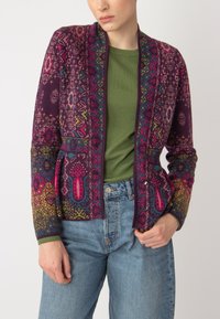 Cardigan tricoté en violet foncé avec des motifs floraux et géométriques multicolores. Il présente un col châle et deux poches à l'avant.