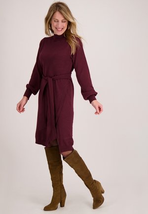 Bordeauxrode jurk met lange mouwen en een tailleceintuur, gemaakt van zachte stof; gecombineerd met bruine suède laarzen tot aan de knie. Getailleerd aan de bovenkant, uitlopend naar de onderkant.