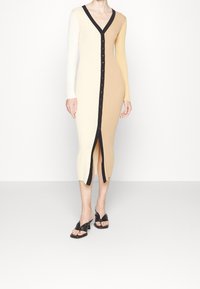 Abito midi a costine in crema e beige, con scollo a V profondo, maniche lunghe, chiusura frontale a bottoni e spacco laterale. Bordo in contrasto nero.