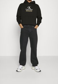 Person som bär en svart Calvin Klein-hoodie, mörka lösa jeans och vita klumpiga sneakers, stående med händerna i fickorna mot en vit bakgrund.