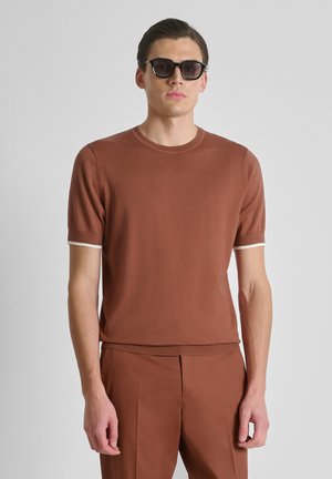 Uomo che indossa un maglione marrone a maniche corte con rifiniture bianche e pantaloni marroni abbinati, in piedi davanti a uno sfondo grigio chiaro, con occhiali da sole neri.