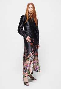 Sort satin kjole med lange puffærmer, prydet med et farverigt blomsterprint i kanten og en tætsiddende talje. Strappy sorte hæle fuldender looket.