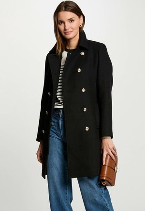 Manteau en laine noir au design structuré, boutons croisés et poches latérales. Jean bleu et sac à main marron.