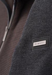 Grauer Reißverschluss-Pullover mit gerippter Textur; verfügt über ein schwarzes Patch mit dem "bugatti"-Logo. Der Hintergrund zeigt braune vertikale Streifen.