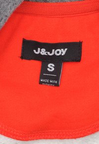 Rode stoffen label met "J&JOY" in witte tekst, maat "S" in zwart, en "MADE WITH DIGNITY" in het wit op een zwart label.