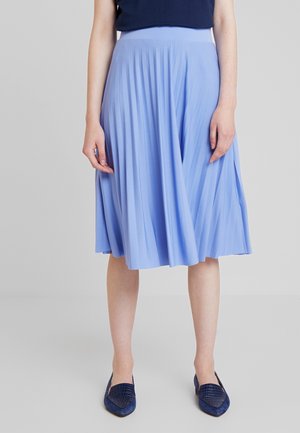 Vrouw draagt een lichtblauwe geplooide rok tot op de knie, een donkerblauw topje en donkerblauwe puntige platte schoenen, staand tegen een effen achtergrond.