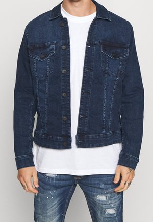 Mann trägt eine dunkelblaue Jeansjacke über einem weißen T-Shirt und zerrissene blaue Jeans, steht vor einem einfarbigen Hintergrund.