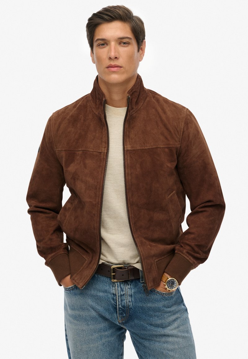 Giacca bomber in suede marrone con collo alto, chiusura con zip e polsini a coste. Indossata sopra una camicia grigia chiara e abbinata a jeans blu.