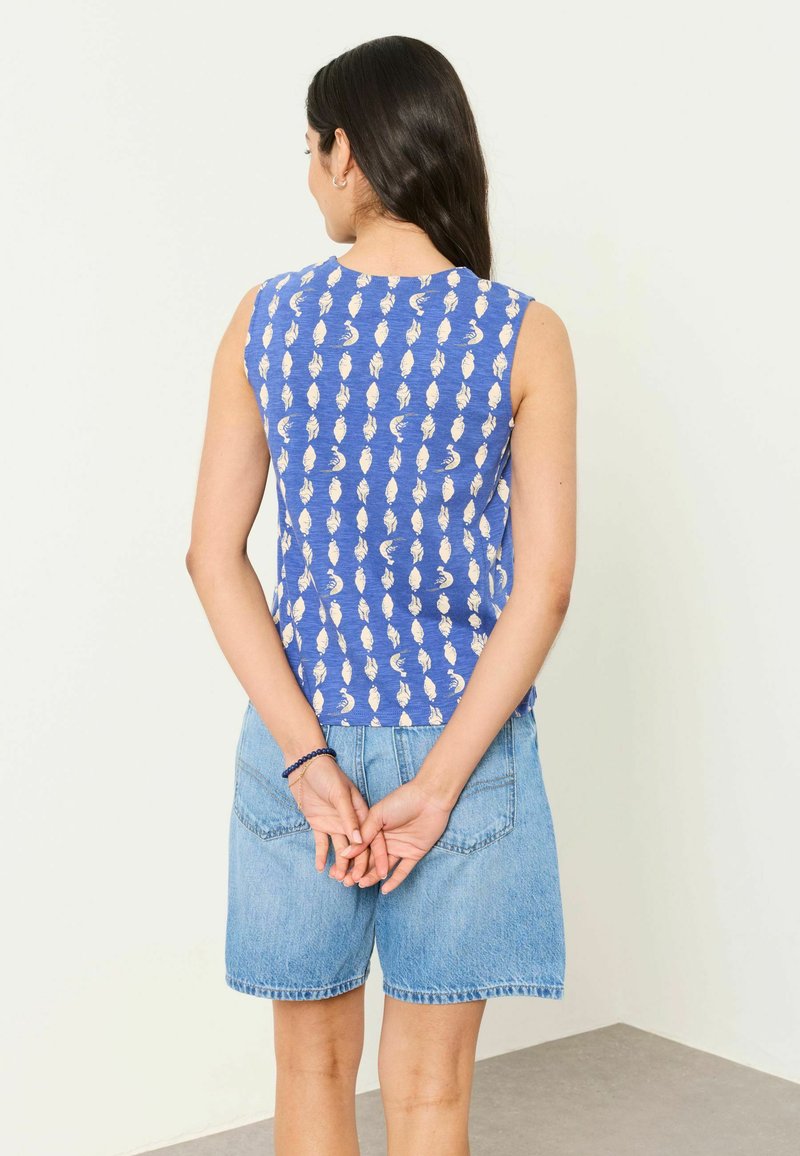 Mujer de pie con las manos entrelazadas detrás de la espalda, vistiendo una camiseta azul sin mangas con un patrón de hojas blancas y pantalones cortos vaqueros.