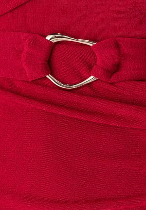 Robe en tissu rouge présentant une surface texturée avec un détail nœud et une boucle métallique en forme de cœur en accent à la taille.