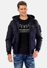 Marineblauer Bomberjacke mit schwarzen Lederelementen, Frontreißverschluss, mehreren Taschen und Kapuze. Getragen über einem schwarzen Grafik-T-Shirt und hellen Jeans.