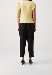 Osoba stojąca tyłem, ubrana w żółtą bluzkę bez rękawów z falbanami, czarne spodnie i czarne buty na obcasie typu kitten heel, na białym tle.